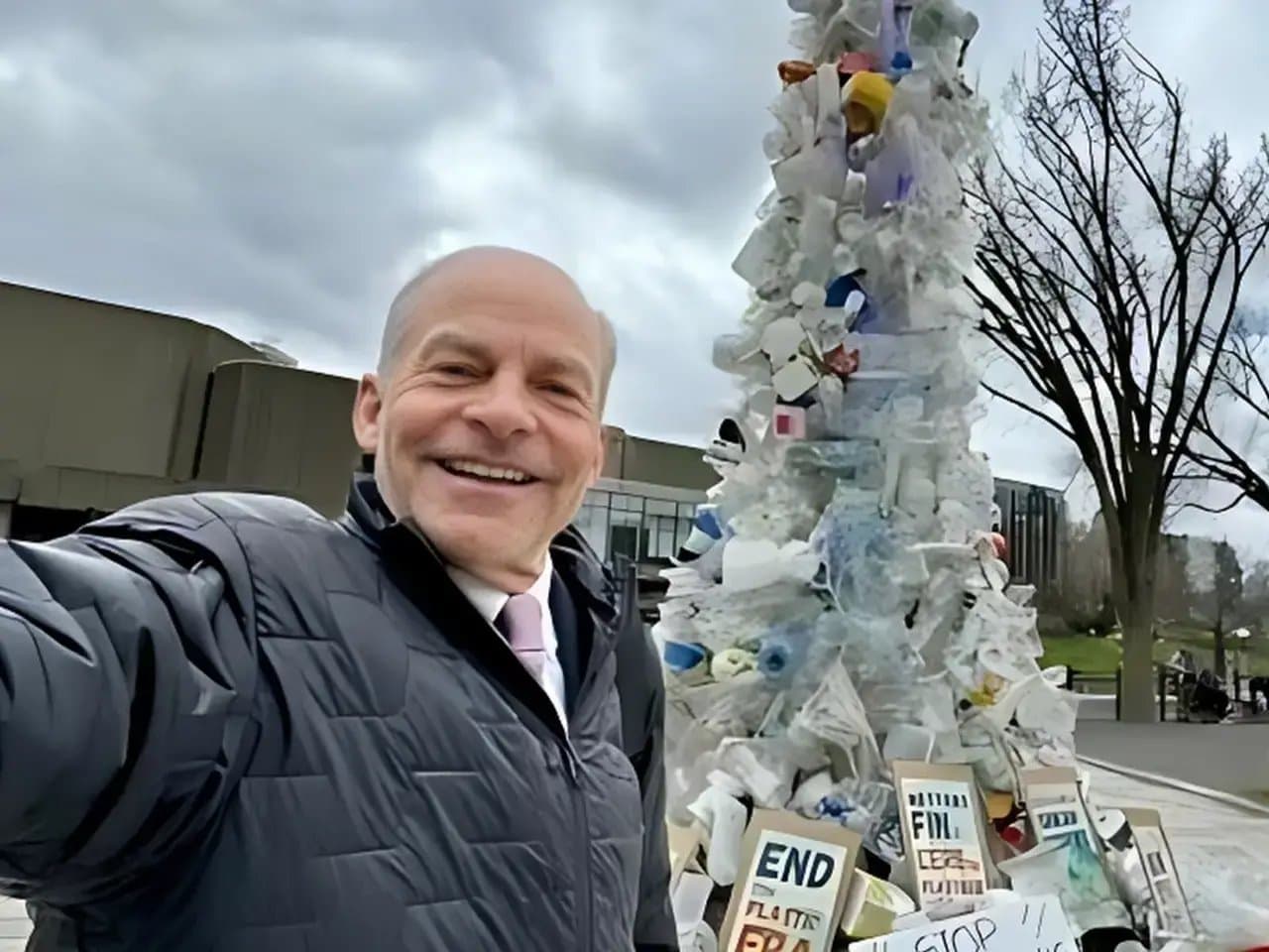 A person smiles in front of a tall sculpture made from plastic waste, with protest signs reading "END PLASTIC" and "STOP" against a backdrop of cloudy skies and bare trees.