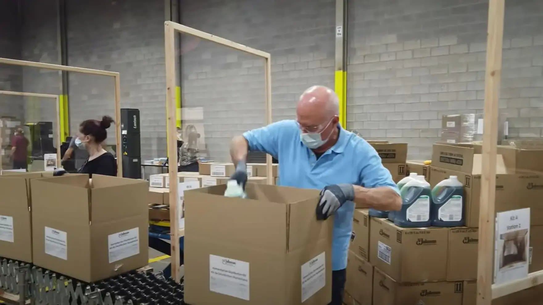 A person wearing a mask and gloves packs items into large cardboard boxes in a warehouse. Text includes: “SC Johnson” and “Kit de Limpieza.” Cleaning products surround him.