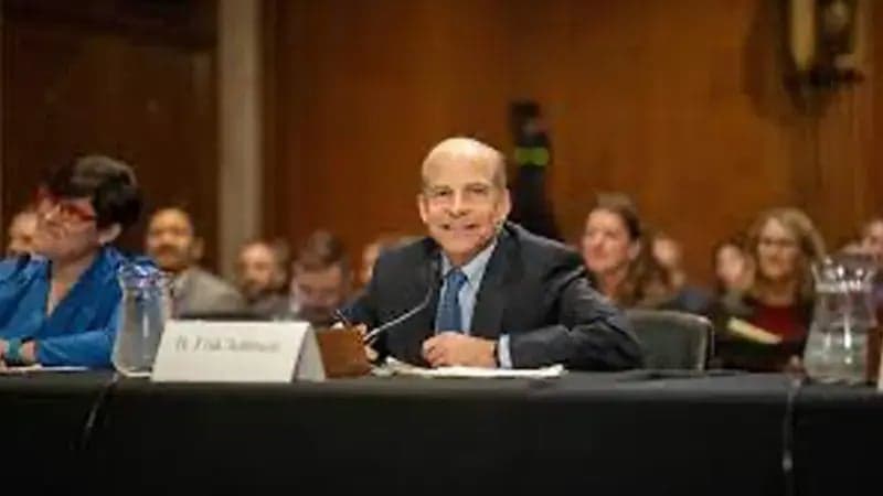 A man sits at a table, speaking into a microphone in a formal hearing room. He is surrounded by others listening. A nameplate on the table reads "H Phil Zamore."