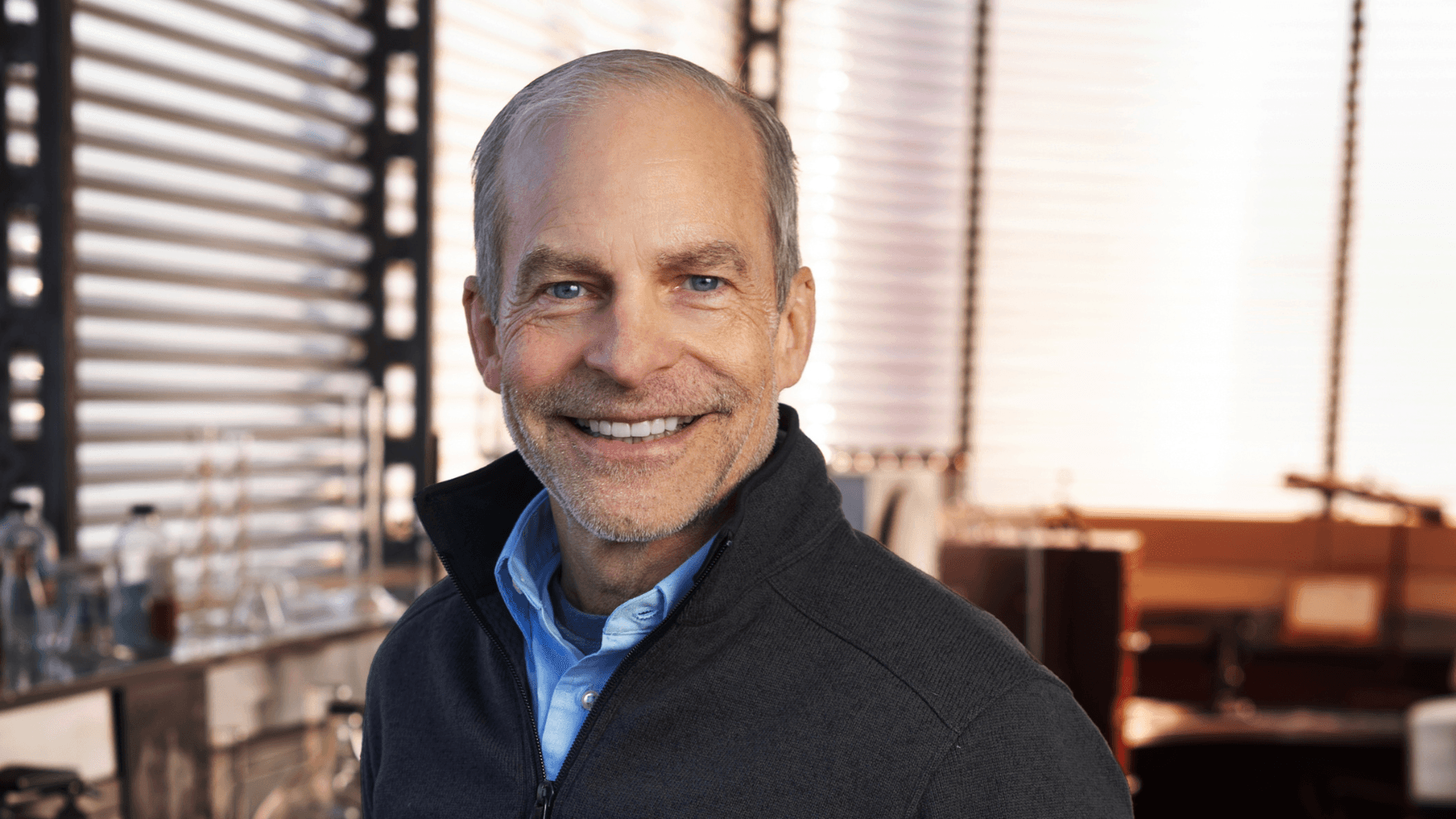 A person is smiling while wearing a light blue shirt and black zipper. He is standing in front of a window with horizontal blinds, with laboratory equipment visible in the background.